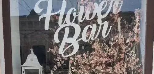 Salona skatlogs ar baltu uzrakstu „Flower Bar” un ziedošiem zariem